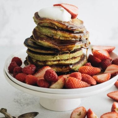 Close up of a stack of matcha pancakes with strawberry topping, whipped cream, and syrup
