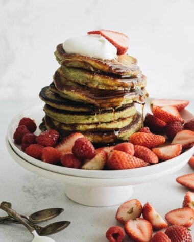 Close up of a stack of matcha pancakes with strawberry topping, whipped cream, and syrup
