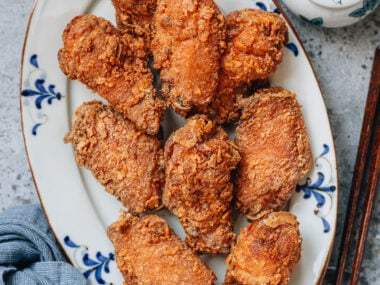 A plate of golden, crispy Chinese fried chicken wings served on a white platter with blue patterns, with a bowl of salt and chopsticks on the side.