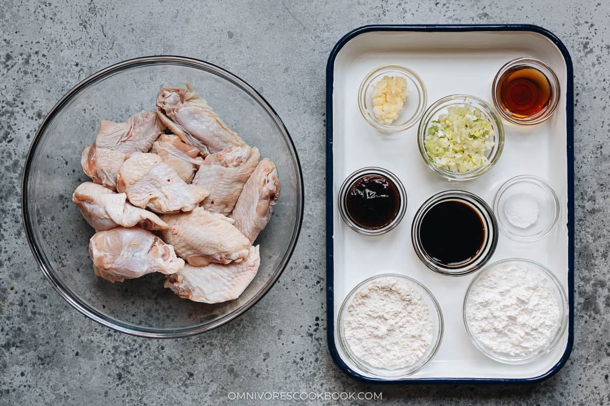 Raw chicken wings in a glass bowl beside small bowls of garlic, scallion, Shaoxing wine, soy sauce, oyster sauce, flour, cornstarch, and salt, all laid out for preparation.