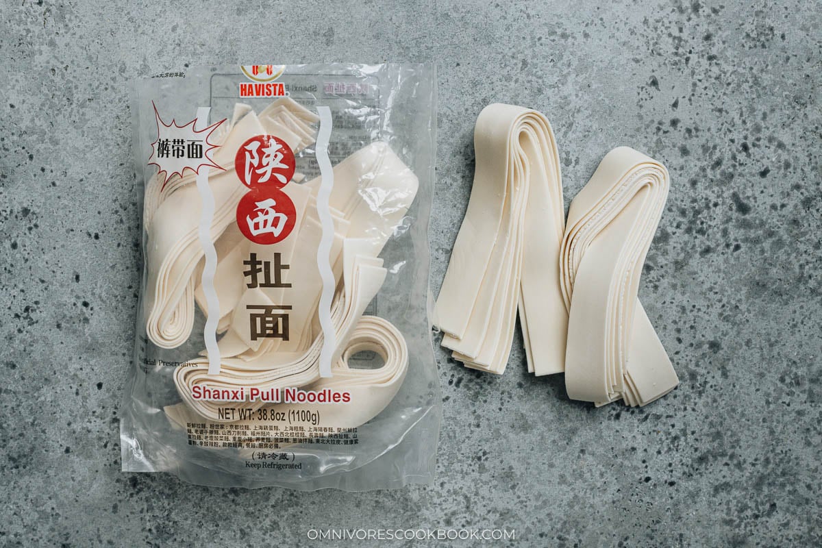 Package of fresh Shanxi pull noodles next to bundles of uncooked wide noodle sheets.
