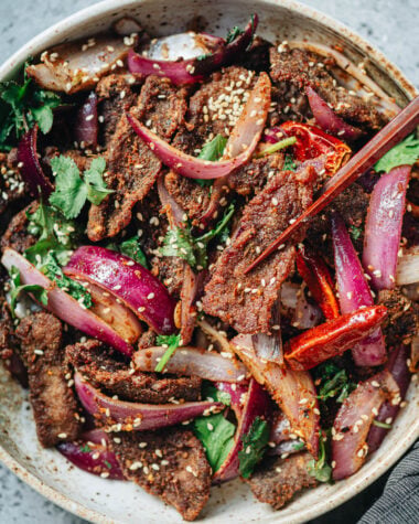 Chopsticks picking up a piece of cumin beef from a bowl filled with red onions and cilantro.