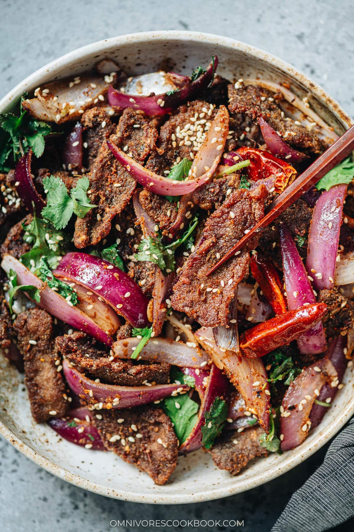 Chopsticks picking up a piece of cumin beef from a bowl filled with red onions and cilantro.