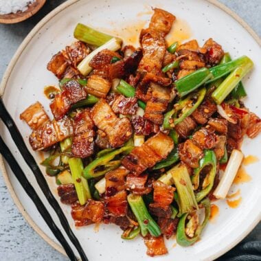 A plate of twice cooked pork with golden pork belly slices, green onions, and green chili peppers tossed in a glossy red sauce, served with black chopsticks.