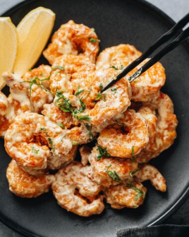 Closeup of a shrimp picked up with black chopsticks, showing the crisp coating and creamy wasabi mayo.
